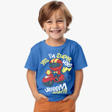 A toddler boy wearing a blue short-sleeve crew neck t-shirt with a superhero graphic on it, standing with hands in pockets and smiling.