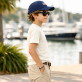 Child wearing a navy cap with an American flag and sunglasses, standing by a waterfront with boats in the background.