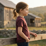 Young girl standing by a wooden fence in a rural setting with a barn and mountains in the background.