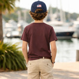 Person wearing a maroon t-shirt and beige pants with a navy cap featuring an American flag, standing by a waterfront with boats.