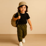 Child wearing a black t-shirt, green pants, and a beige bucket hat with a backpack on a beige background